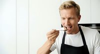 Young Caucasian male chef in black apron tasting food with wooden spoon, smiling while cooking in kitchen. Suitable for culinary, restaurant, cooking class, food blog, and chef profession content.