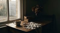 Cozy autumn morning scene with coffee cup, open book and dried flowers on wooden table by window creating warm atmospheric mood for relaxation.