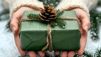 Hands holding green wrapped Christmas gift with pine cone and evergreen branch decoration for holiday season celebrations.