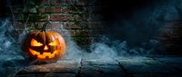 Carved jack-o-lantern pumpkin with glowing face surrounded by blue fog and smoke against weathered brick wall and wooden log for autumn holiday celebrations.