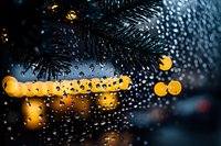Raindrops on window glass with blurred yellow city lights and pine branches creating atmospheric bokeh effect in dark evening, moody winter atmosphere.