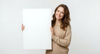 Young Caucasian woman in beige sweater holding blank white poster board with cheerful smile, standing against neutral background for marketing presentations.
