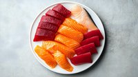 Fresh raw salmon and tuna sashimi slices arranged on white ceramic plate against light textured background, traditional Japanese cuisine preparation.
