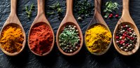 Colorful spices and herbs in wooden spoons on dark background - turmeric, paprika, dried parsley, curry powder and peppercorns arranged in row.