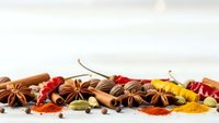 Aromatic spices and herbs scattered on white surface including star anise, cinnamon sticks, cardamom pods, chili peppers and turmeric powder for cooking.