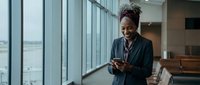 Professional African American businesswoman using smartphone in modern office corridor with large windows, corporate executive checking messages.