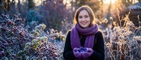 Young Caucasian woman in purple scarf holding fresh blueberries in cupped hands during golden hour sunset with frost-covered plants in background.