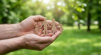 Hands holding wooden family figures in cupped palms outdoors with green nature background symbolizing protection, care and family unity concept.