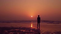 Silhouette of young man standing alone on beach at sunset with reflection on wet sand, dramatic orange sky and sun creating peaceful solitary moment.