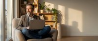 Mature businessman working on laptop from home office, sitting comfortably in armchair with warm natural lighting and modern interior background.