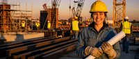 Smiling female construction engineer in yellow hard hat holding blueprints at busy building site during golden hour sunset with cranes and workers in background.