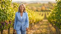 Professional middle-aged Caucasian businesswoman in light blue suit standing confidently in vineyard rows during golden hour sunset for corporate lifestyle.
