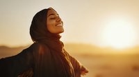 Young Middle Eastern woman in hijab smiling with eyes closed against golden sunset backdrop, arms outstretched embracing nature and freedom.