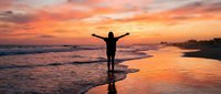 Person with raised arms celebrating freedom on beach at golden sunset with waves and dramatic orange sky reflecting on wet sand for wellness concepts.