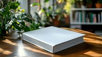 White hardcover book mockup on wooden table with natural sunlight and green plants, blank template for branding and design presentation in cozy home office setting.
