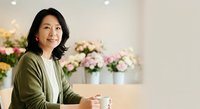Middle-aged Asian woman in olive cardigan and white shirt sitting indoors with blurred floral arrangements in background, suitable for business, lifestyle, and corporate communications.