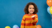 Happy mixed-race girl with curly hair wearing colorful striped sweater pointing at camera on blue background with balloons for birthday party celebration.