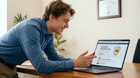 Smiling Caucasian man in blue sweater using laptop computer for online security protection at home office desk with modern interior background.