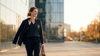 Professional businesswoman walking confidently in modern urban district during golden hour sunset, carrying leather bag and blazer for corporate lifestyle.