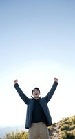 Young man of East Asian descent wearing a dark blue jacket and beige pants celebrates success with arms raised against clear sky, standing on hillside with golden vegetation below, expressing joy and achievement.