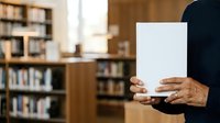 Person holding blank white book or magazine in library with wooden shelves and books in background for publishing and education concepts.