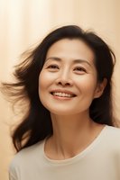Asian woman with flowing black hair and genuine smile wearing casual white top against warm beige background, expressing joy and confidence in natural lighting.
