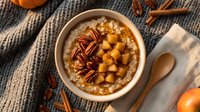 Creamy oatmeal bowl topped with roasted chickpeas, pecans, and cinnamon sticks on gray knit fabric with wooden spoon and pumpkin.