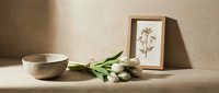 Minimalist still life with white tulips, ceramic bowl, and botanical framed print on beige textured surface in warm natural light.