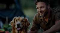 Caucasian bearded man in brown t-shirt smiling with friendly golden retriever dog outdoors. Natural lighting and bokeh background create warm emotional connection.