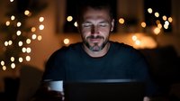 Caucasian man with beard working late at night on laptop, illuminated by screen glow with bokeh lights in background, expressing contentment while drinking coffee.