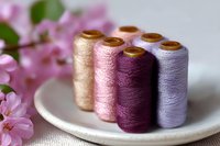 Colorful sewing thread spools in pastel purple, pink, and gold tones arranged on white ceramic plate with soft focus cherry blossoms in background.