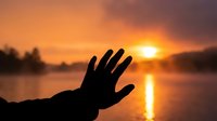 Hand silhouette reaching toward golden sunrise over calm lake water with misty atmospheric lighting creating peaceful morning meditation scene.