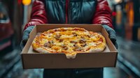 Fresh hot pizza with melted cheese and toppings in delivery box held by courier wearing winter gloves and red jacket on blurred urban background.