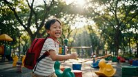Young Asian girl with backpack laughing joyfully at colorful playground during golden hour sunlight filtering through trees creating warm outdoor recreation scene.