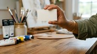Mature male artist holding blank white card at wooden desk with paintbrushes, markers and supplies, suitable for creative business, art studio, and portfolio presentation concepts.