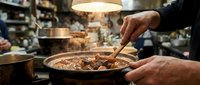 Skilled male chef stirring a steaming clay pot of braised meat in a busy restaurant kitchen under warm lamp light.