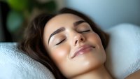 Young Mediterranean woman with closed eyes and serene expression lying on white pillow, glowing healthy skin and natural makeup for wellness and beauty concepts.
