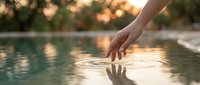 Woman hand gently touching calm water surface creating ripples at sunset with warm golden light reflecting on pool water for relaxation and wellness concepts.
