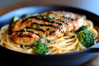 Grilled chicken breast with spaghetti pasta and steamed broccoli in black ceramic bowl, garnished with fresh herbs and black pepper, close up view.