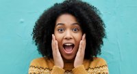 Excited young African American woman with curly hair showing surprise expression with hands on face against turquoise wall for lifestyle marketing.