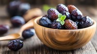 Fresh ripe plums in wooden bowl with mint leaf garnish on rustic table, selective focus on dark purple fruits, natural food photography.