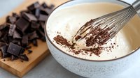 Creamy white mixture in ceramic bowl with chocolate being whisked in, dark chocolate baking preparation.
