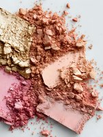 Crushed makeup powders in gold, coral, and pink tones creating a luxurious palette of shimmering cosmetic textures on white background.