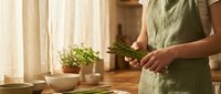 Woman in green apron preparing fresh herbs in modern kitchen with natural lighting and wooden countertop for healthy cooking and meal preparation.