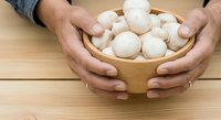 Hands holding wooden bowl filled with fresh white mushrooms on natural wood surface for cooking and food preparation.