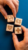Hands holding wooden blocks with engraved healthcare symbols including stethoscope, heartbeat, medical cross and dollar sign on blue background for insurance and medical cost concepts.