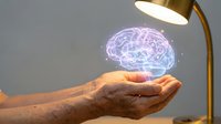 Mature adult hand holding glowing purple holographic brain with desk lamp in background, representing artificial intelligence, innovation, and digital technology concepts.
