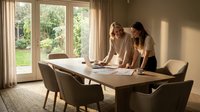 Two professional businesswomen collaborating over documents at modern conference table in bright office with garden view during productive meeting.