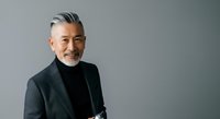 Mature Asian man with gray hair and beard wearing black turtleneck and charcoal blazer, smiling at camera against neutral gray background for corporate portraits and business headshots.