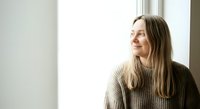 Young woman with long brown hair and fair complexion wearing a beige knit sweater, gazing thoughtfully toward window light in minimalist interior space.